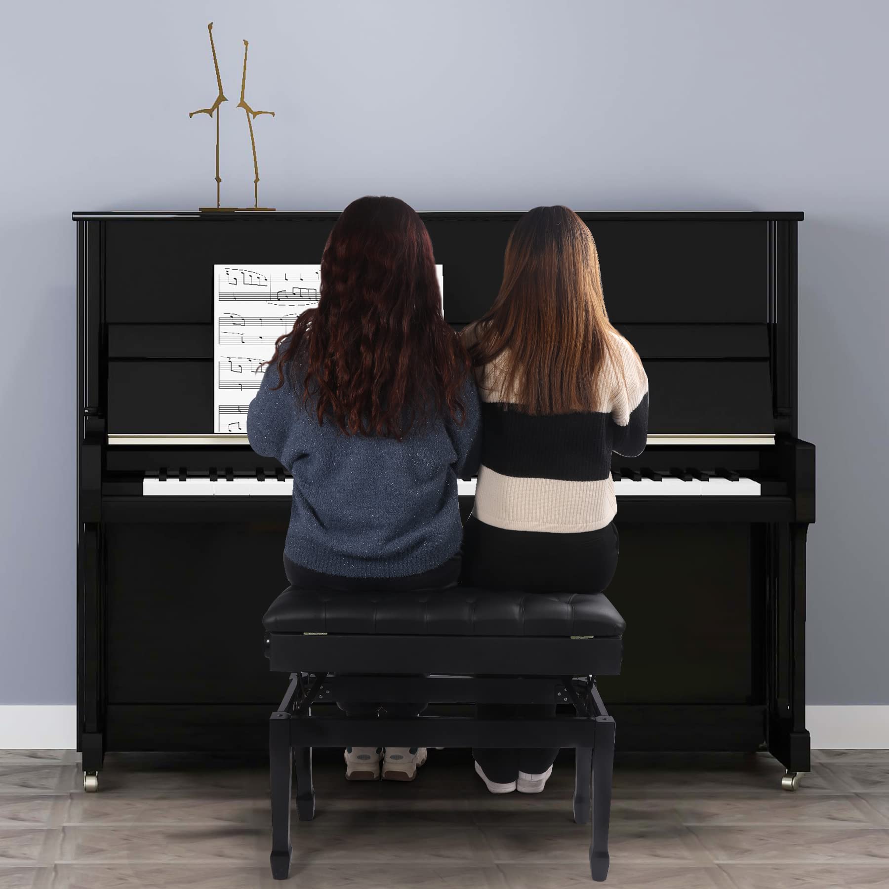 Two people playing a black upright piano against a gray wall.